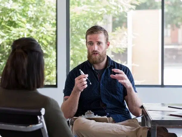 A bearded man sitting at a table talking to a woman, holding a pen